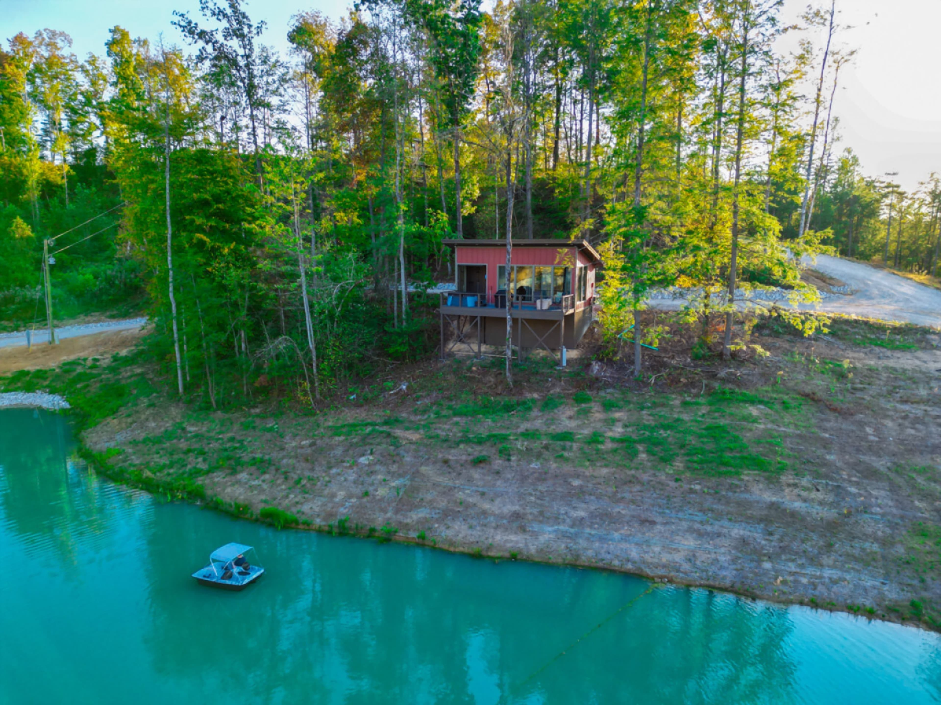 Aerial view of a red cabin with a deck above a turquoise lake, with a pedal boat on the water