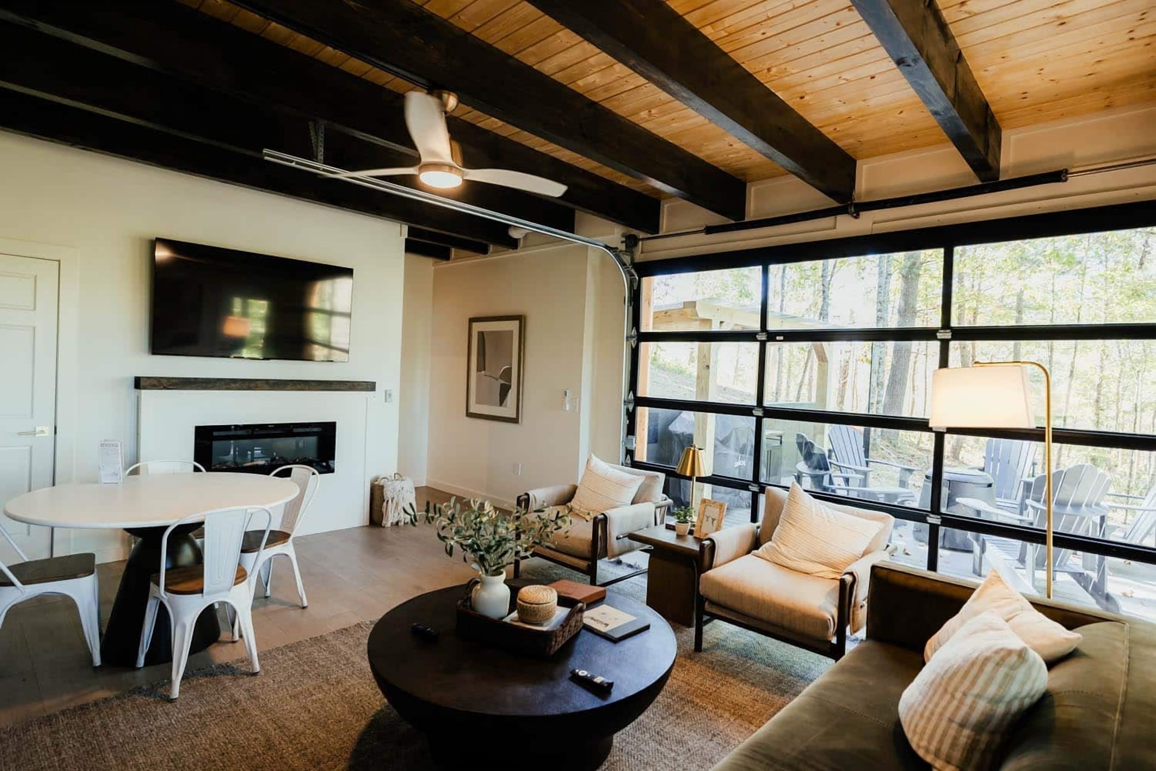 Bright cabin living room with dark ceiling beams, a fireplace, a round dining table and a glass garage door to the deck