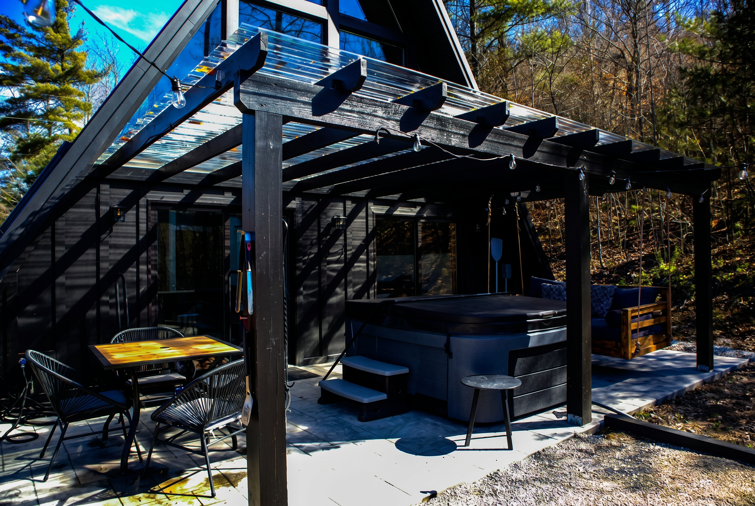 Pergola-covered patio with a hot tub, an outdoor dining table and a porch swing beside a dark A-frame cabin