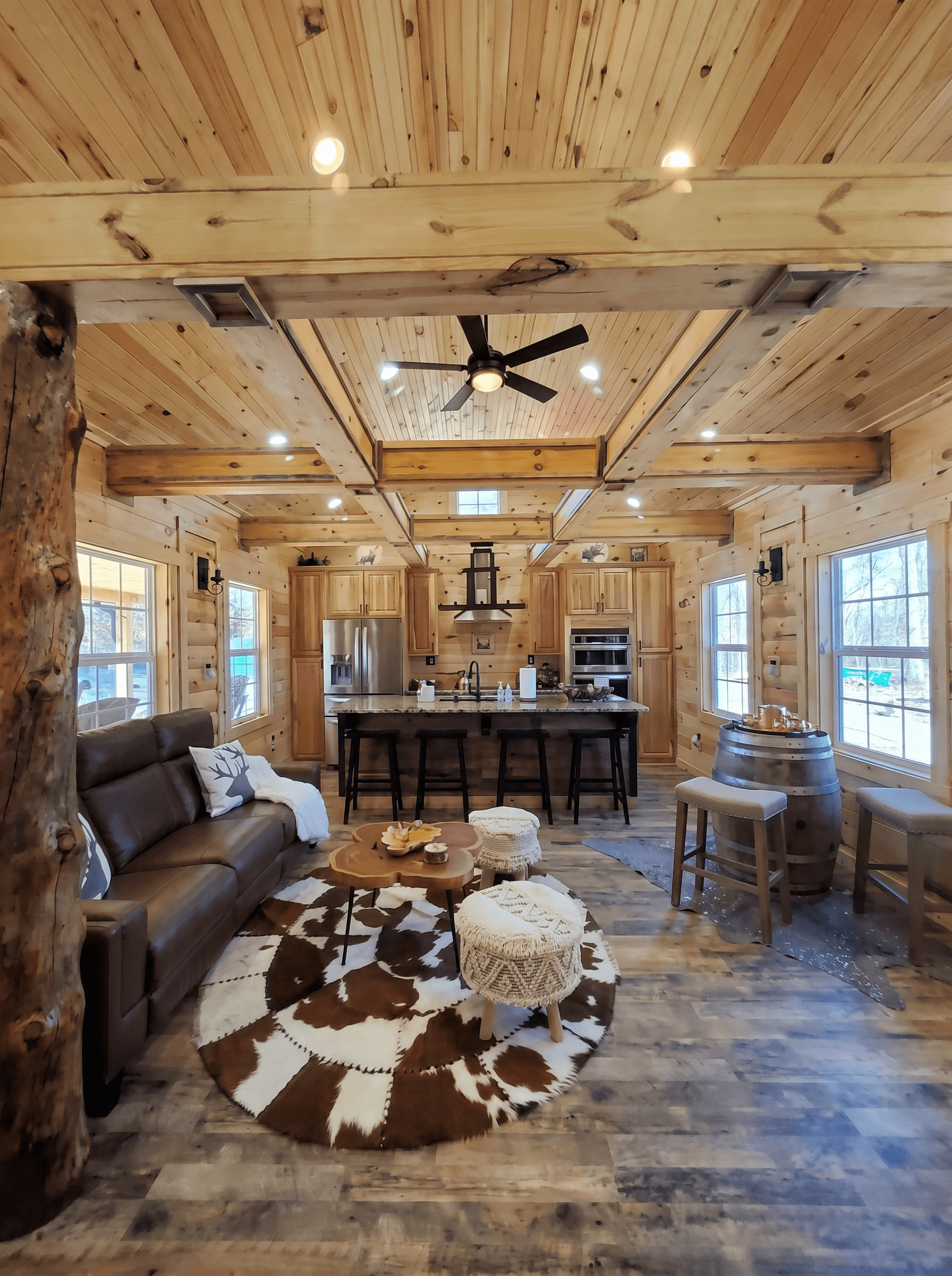 Open cabin living room and kitchen with knotty pine walls and beams, a leather sofa, a cowhide rug and bar stools