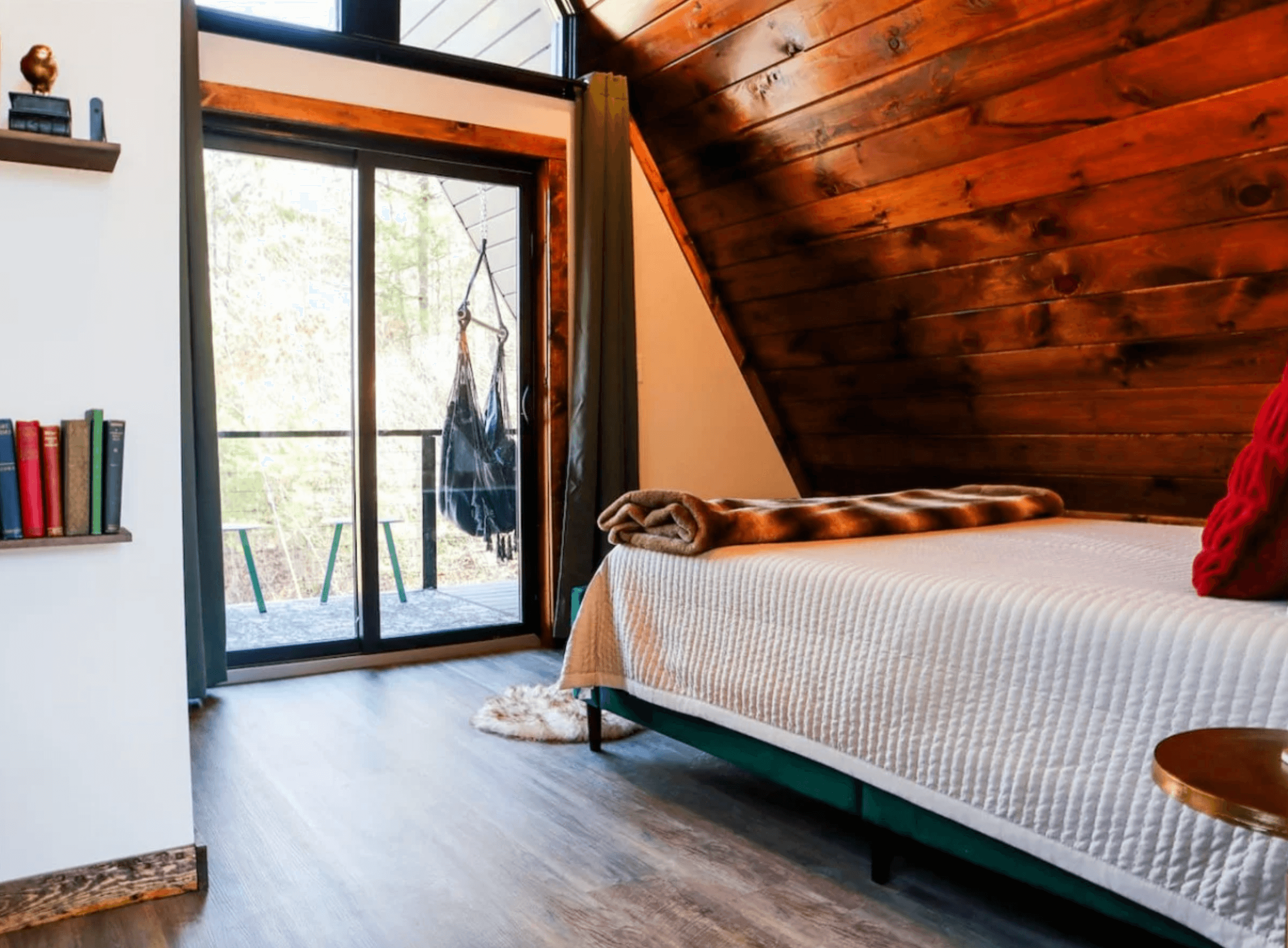 A-frame bedroom with a wood plank ceiling, a made bed and a glass door to a deck with a hanging chair