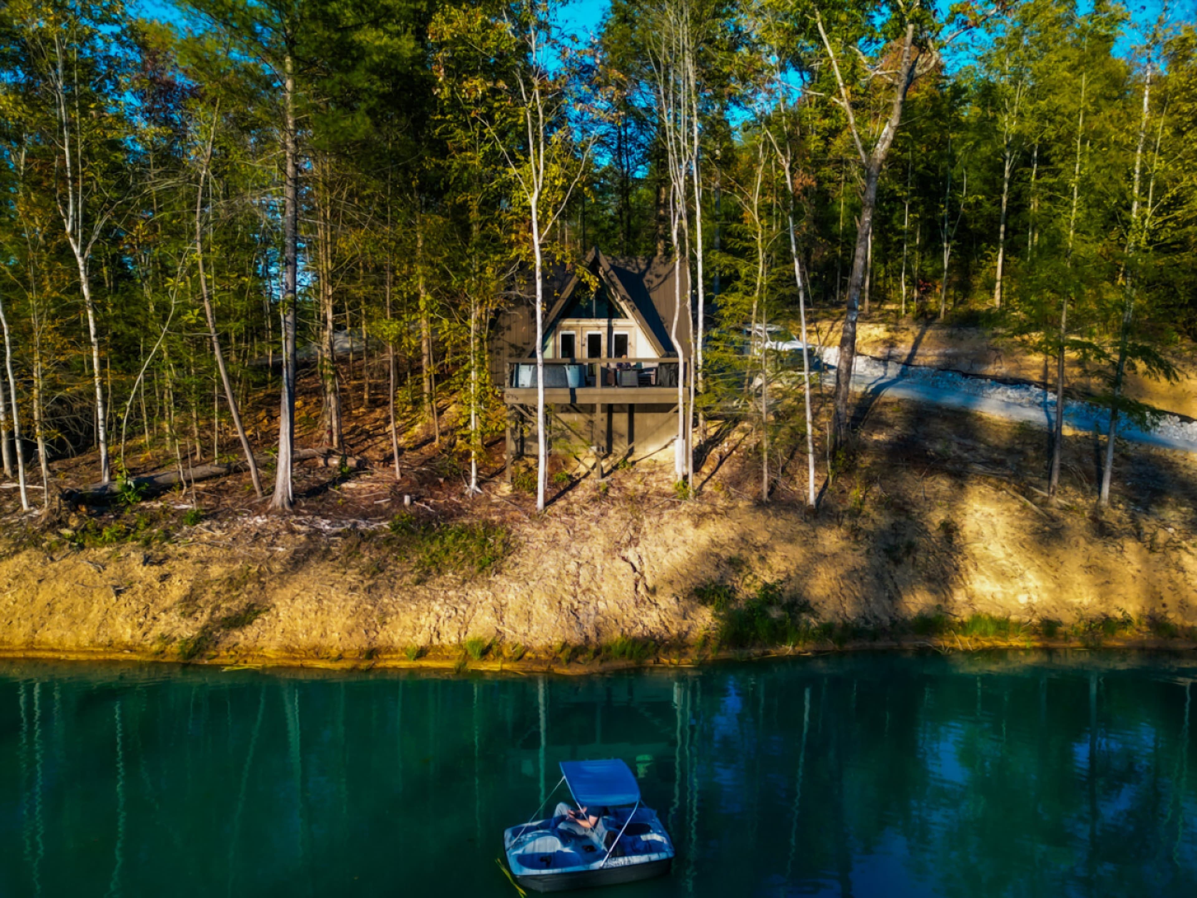 A-frame cabin on a wooded hillside above a lake, with a pedal boat on the water