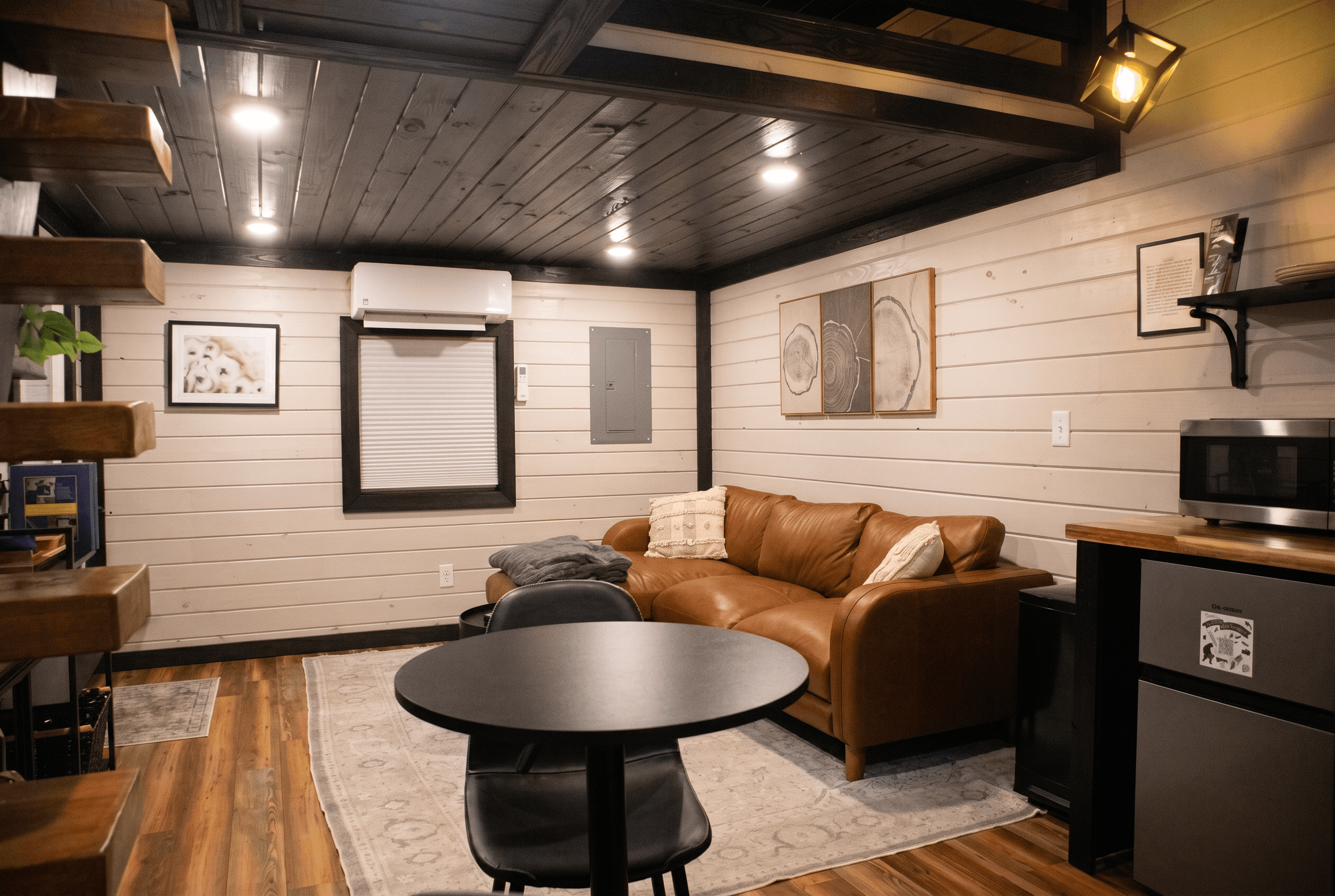 Cabin studio with a tan leather sofa, a small round table, white shiplap walls and a dark wood ceiling