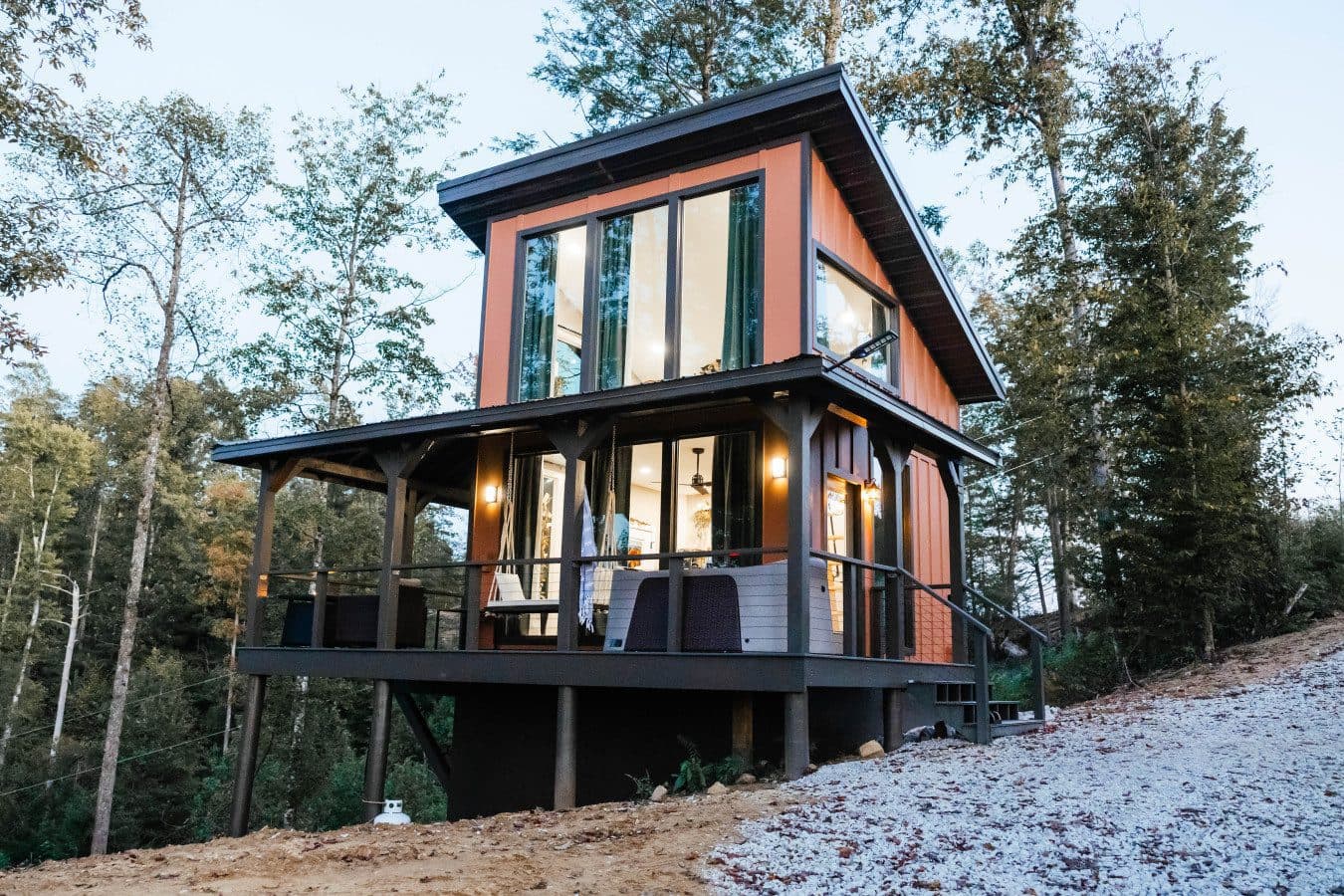 Modern two-story cabin with copper-colored siding, tall windows and a covered porch on a wooded hillside