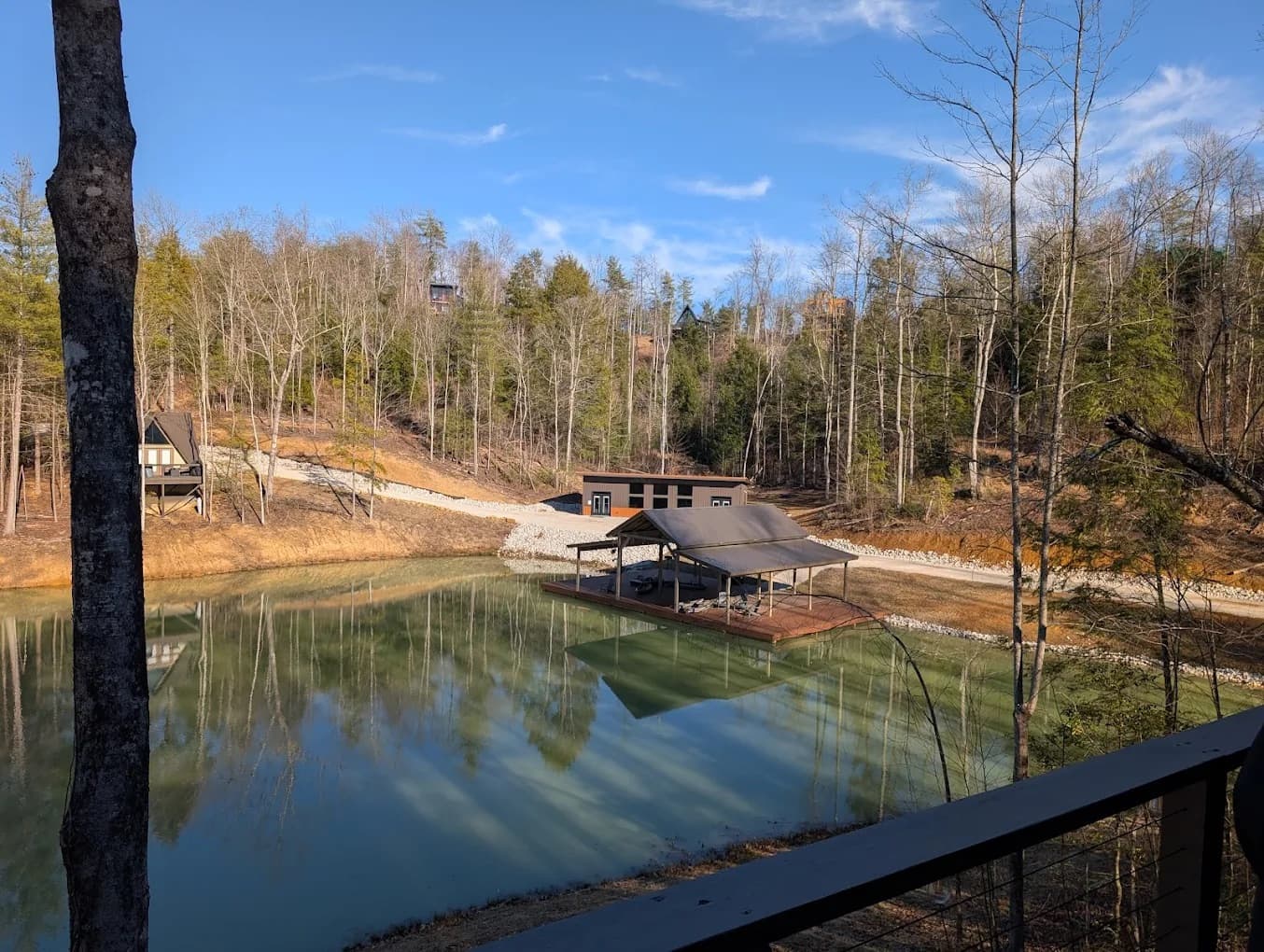 Pond with a covered dock and pavilion, surrounded by forest and cabins, seen from a deck