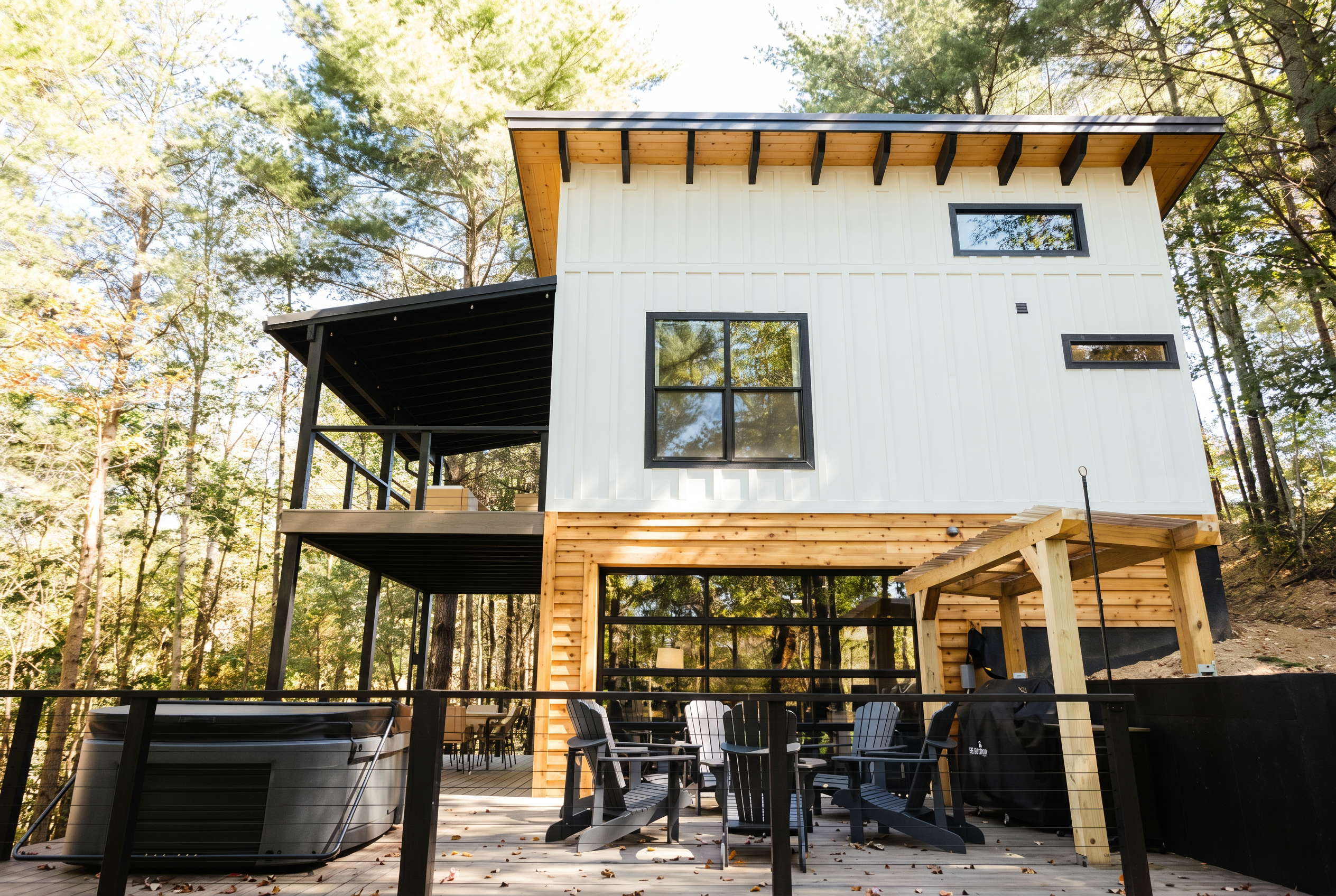 Modern white and cedar cabin with a deck, hot tub, Adirondack chairs and a wooden pergola among tall trees